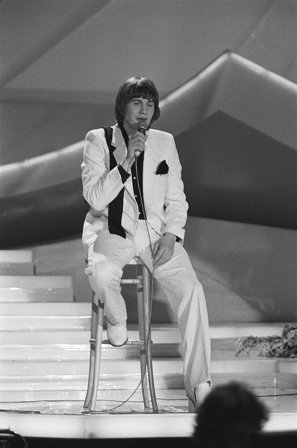 Johnny Logan im weißen Anzug singt auf einem Barhocker sitzend beim ESC 1980 in Den Haag