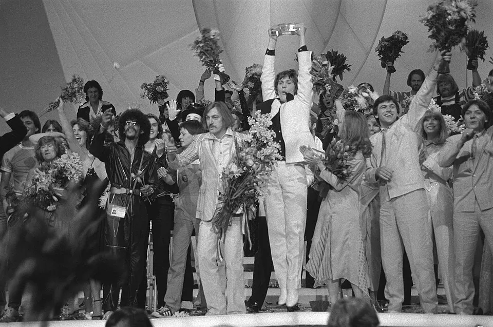 Johnny Logan reckt auf der Bühne die Trophäe hoch, umringt von jubelnden Teilnehmern, ESC 1980