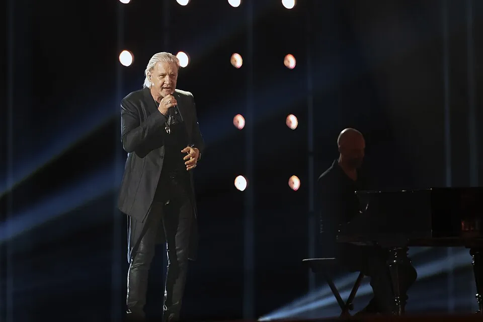 Johnny Logan singt im schwarzen Anzug im Scheinwerferlicht auf der ESC-Bühne 2024, daneben ein Pianist