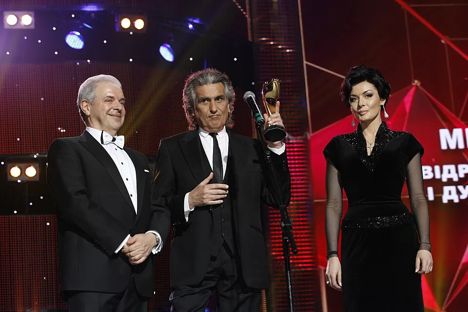 Toto Cutugno mit Preisstatue und Mikrofon auf einer Bühne, neben ihm ein Mann im Smoking und eine Frau, 2013