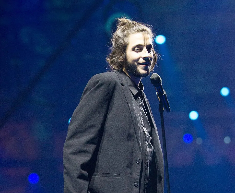 Salvador Sobral im dunklen Anzug lächelt am Mikrofon auf blau beleuchteter Bühne, ESC 2017 in Kiew